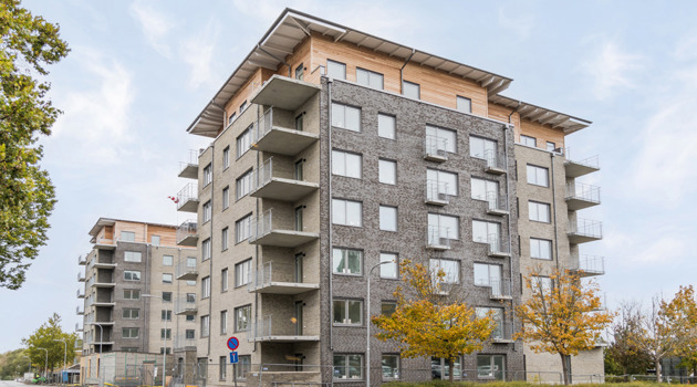 Flerbostadshus i grått tegel med balkonger och träfasad högst upp, sett från gatan med höstfärgade träd framför.