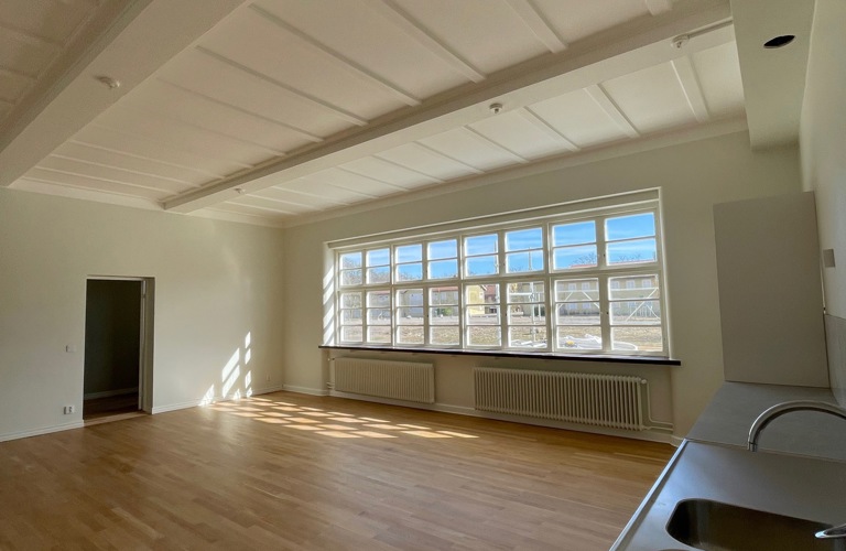 Bild på öppen planlösning mellan kök och vardagsrum med vita väggar och trägolv. Stort fönsterparti längs med hela väggen.