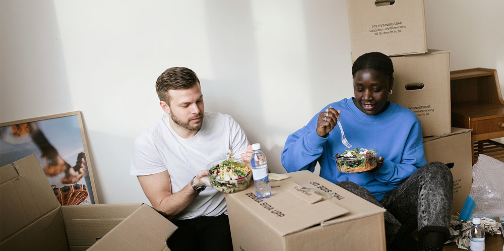 En man och en kvinna som sitter bland flyttkartonger och äter sallad