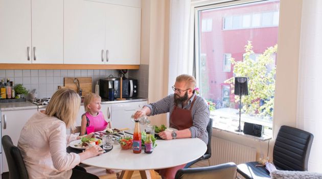 Vuxna och barn sitter runt ett matbord och äter tillsammans i köket.
